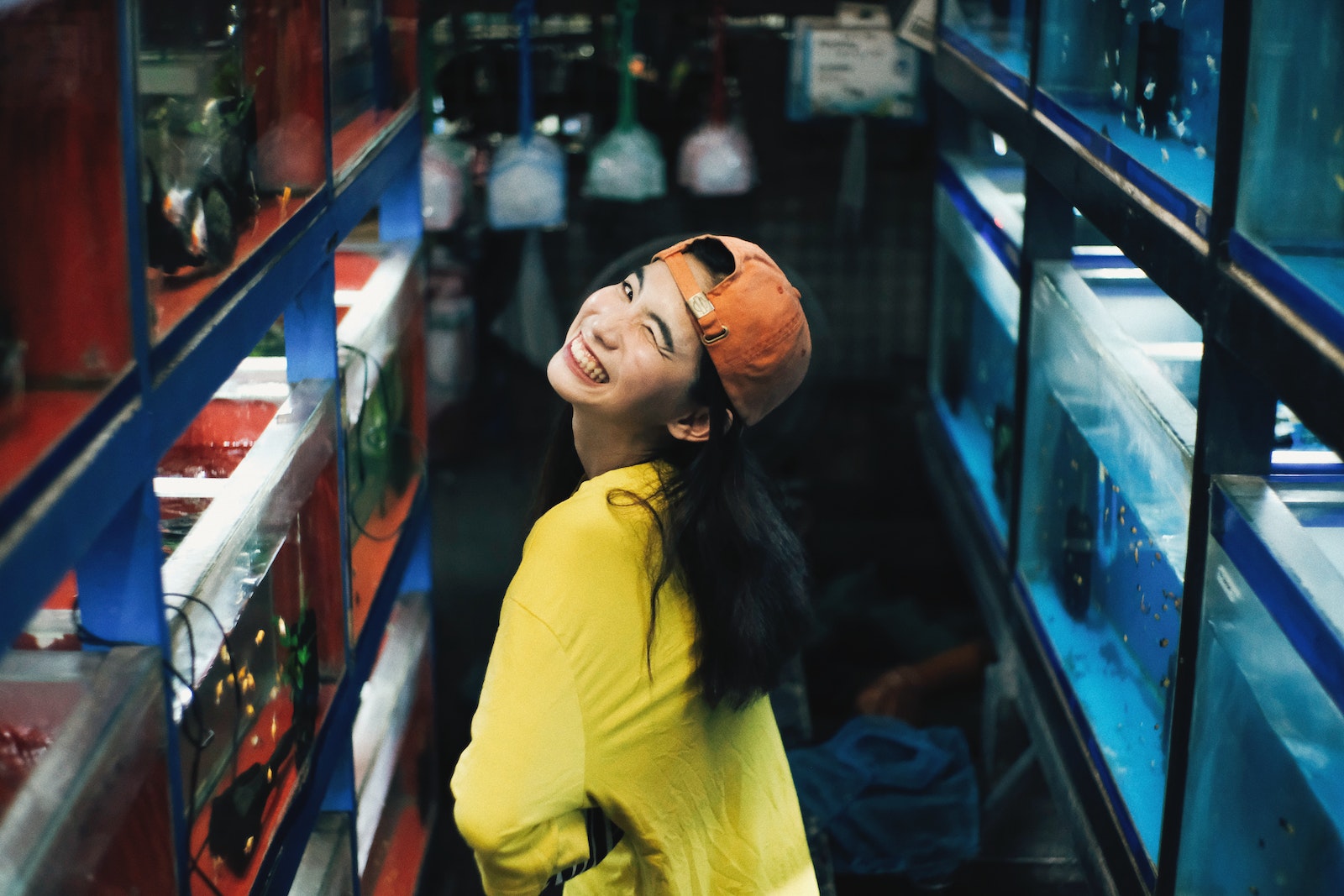 Photo of Smiling Woman in Yellow Top and Orange Hat Posing by Fish Tanks