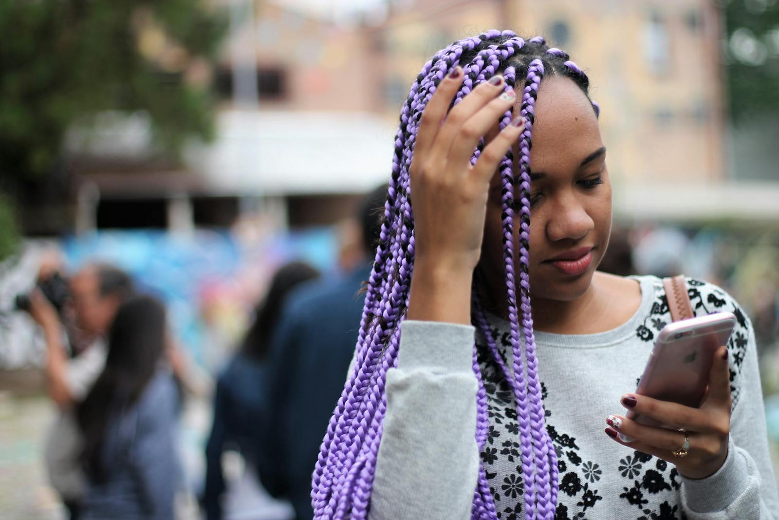 A young woman with vibrant purple braids checks her smartphone in an urban outdoor setting.