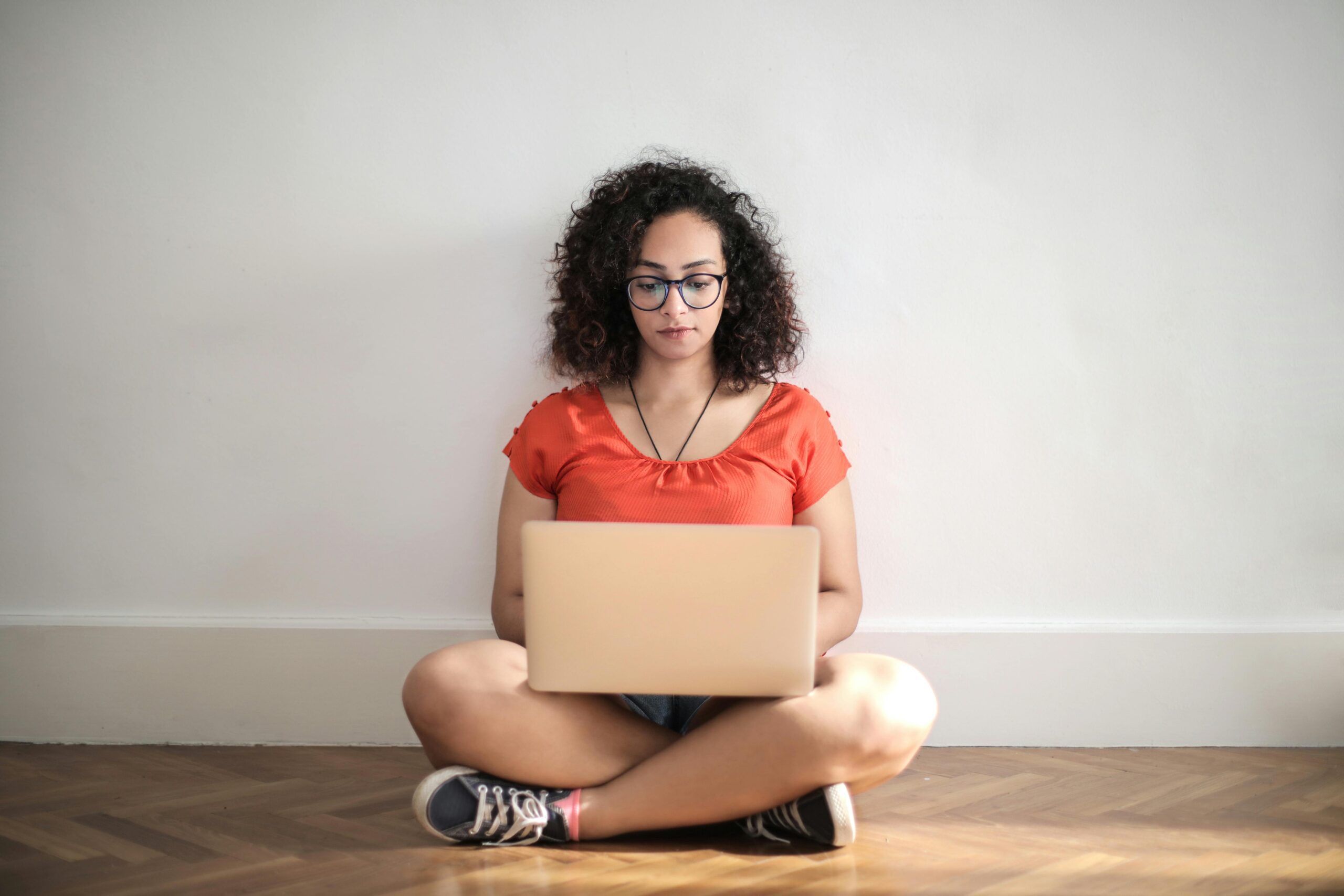 Woman in orange crew neck t shirt sitting on brown wooden floor using laptop